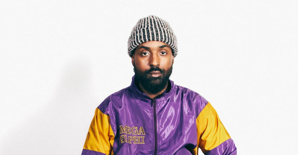 Person wearing a purple and yellow jacket sitting on a stool against a white background